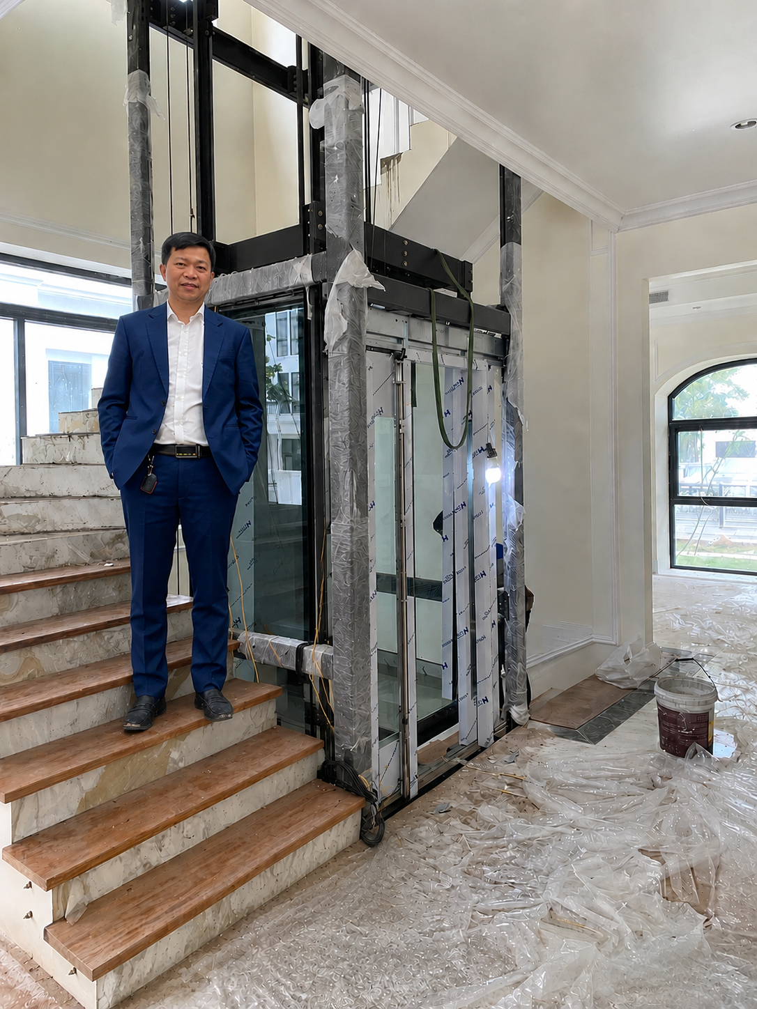 Thang máy gia đình đang lắp đặt cạnh cầu thang gỗ trong nhà, kỹ sư mặc vest xanh đứng bên cạnh